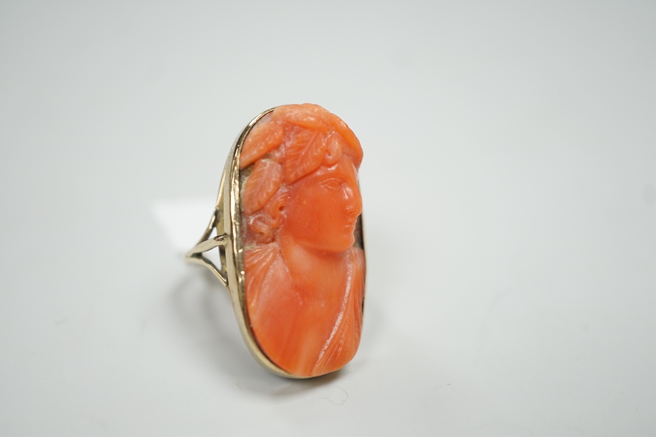 A yellow metal and coral set oval dress upfinger ring, the coral carved with a classical bust to sinister, size O, gross weight 8.2 grams.